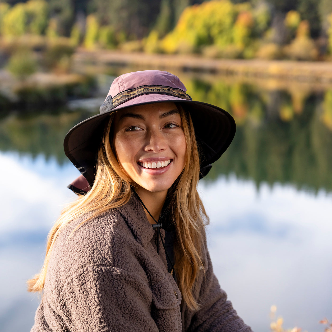 A woman smiling outdoors by a lake, wearing a wide-brimmed dusty purple hat with a dark band and a brown fleece jacket.